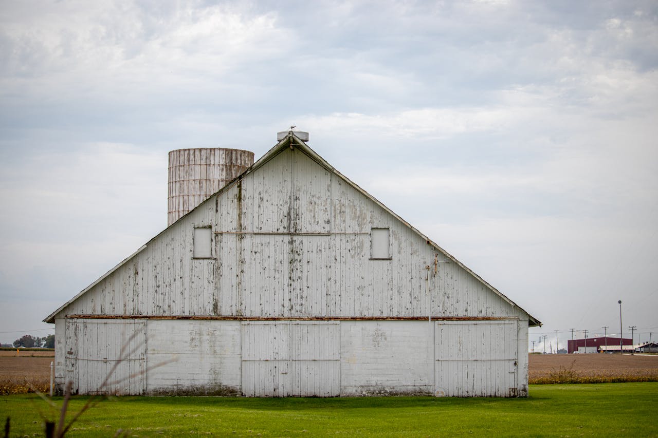 Rustic white barn with silo on a green field under an overcast sky in a rural area.
